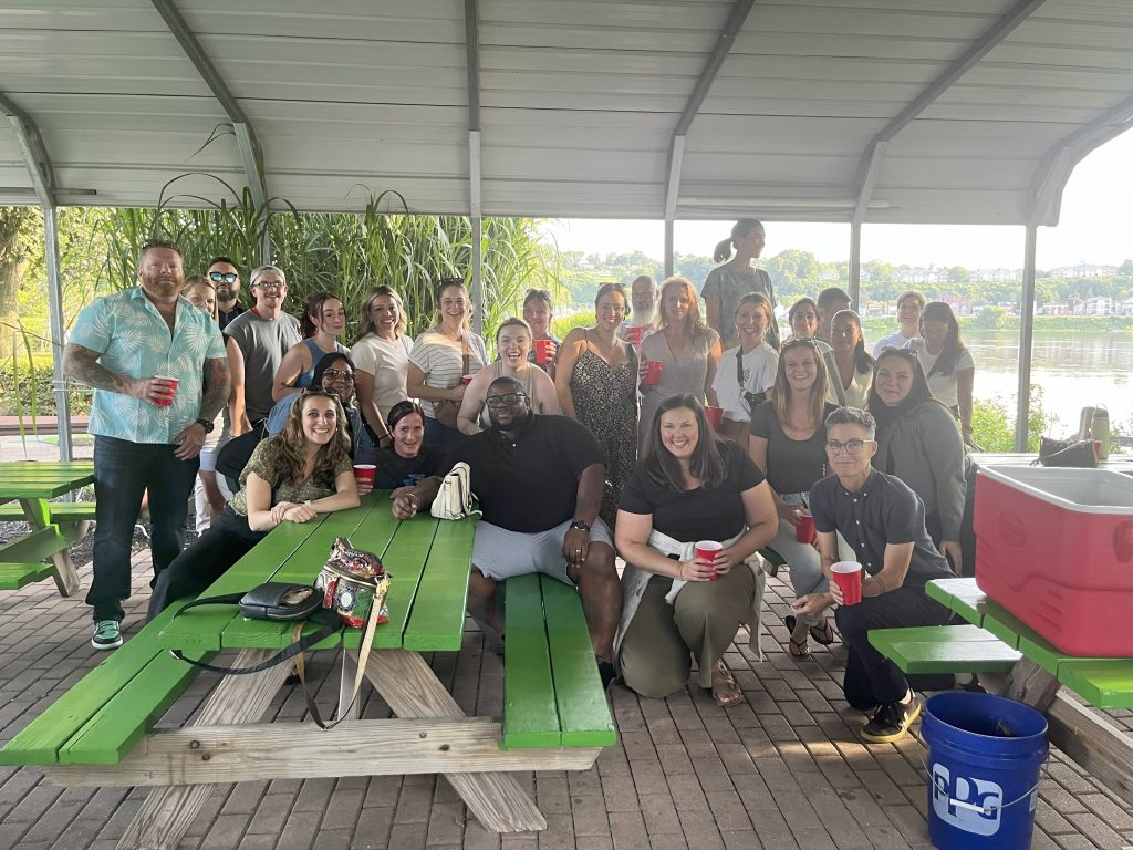 A large group of SWIM staff (Social Workers/social service advocates, Investigators, Mitigators) posing for a group photo around green picnic tables.