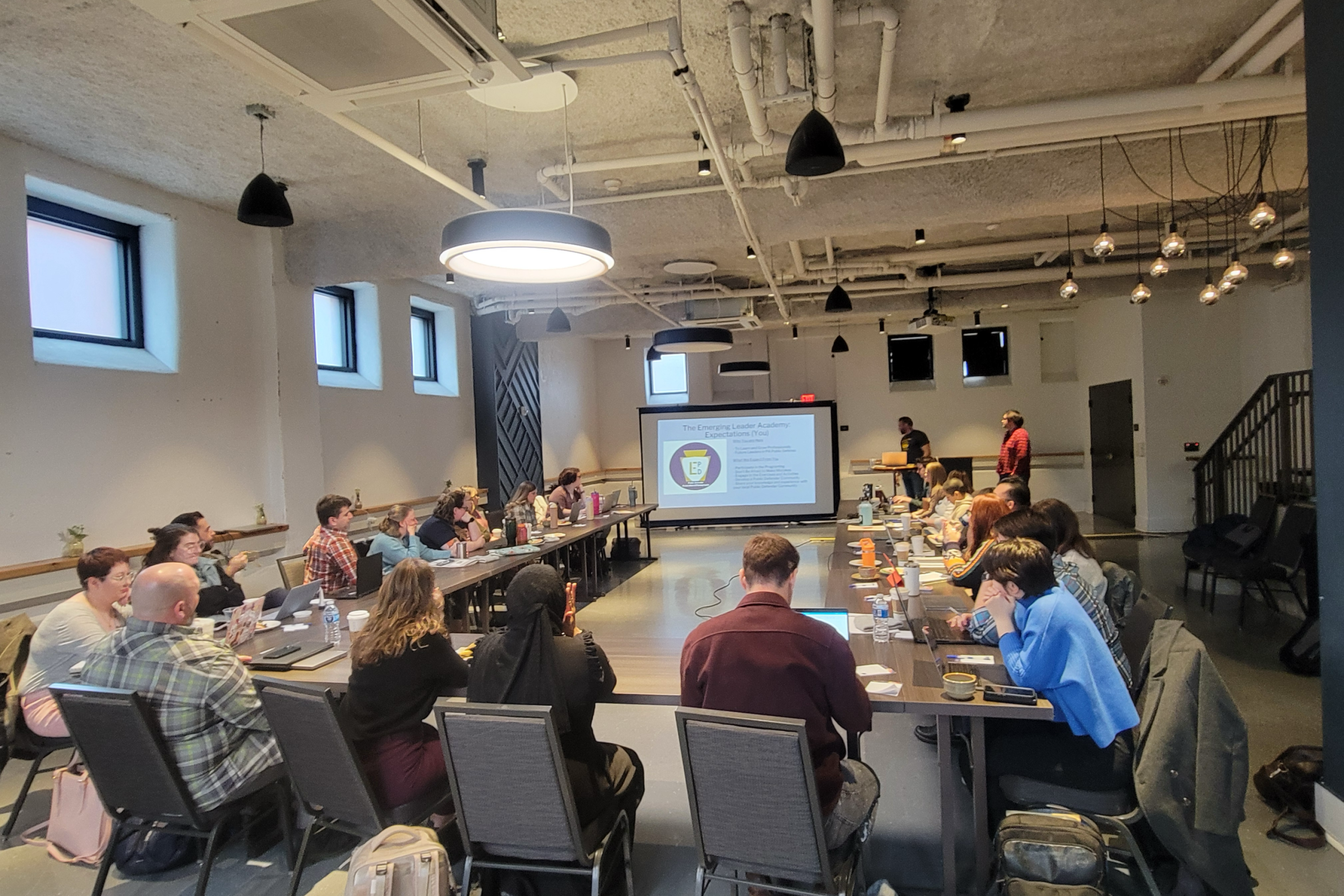 A wide shot of Emerging Leaders, Mentors, and PDAP staff, gathered around a conference room table and looking at a presentation slide reading "Emerging Leader Academy: Expectations (You)".